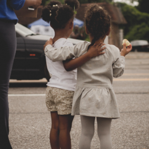 Two young girls watch and wave as cars go by on W. Florissant